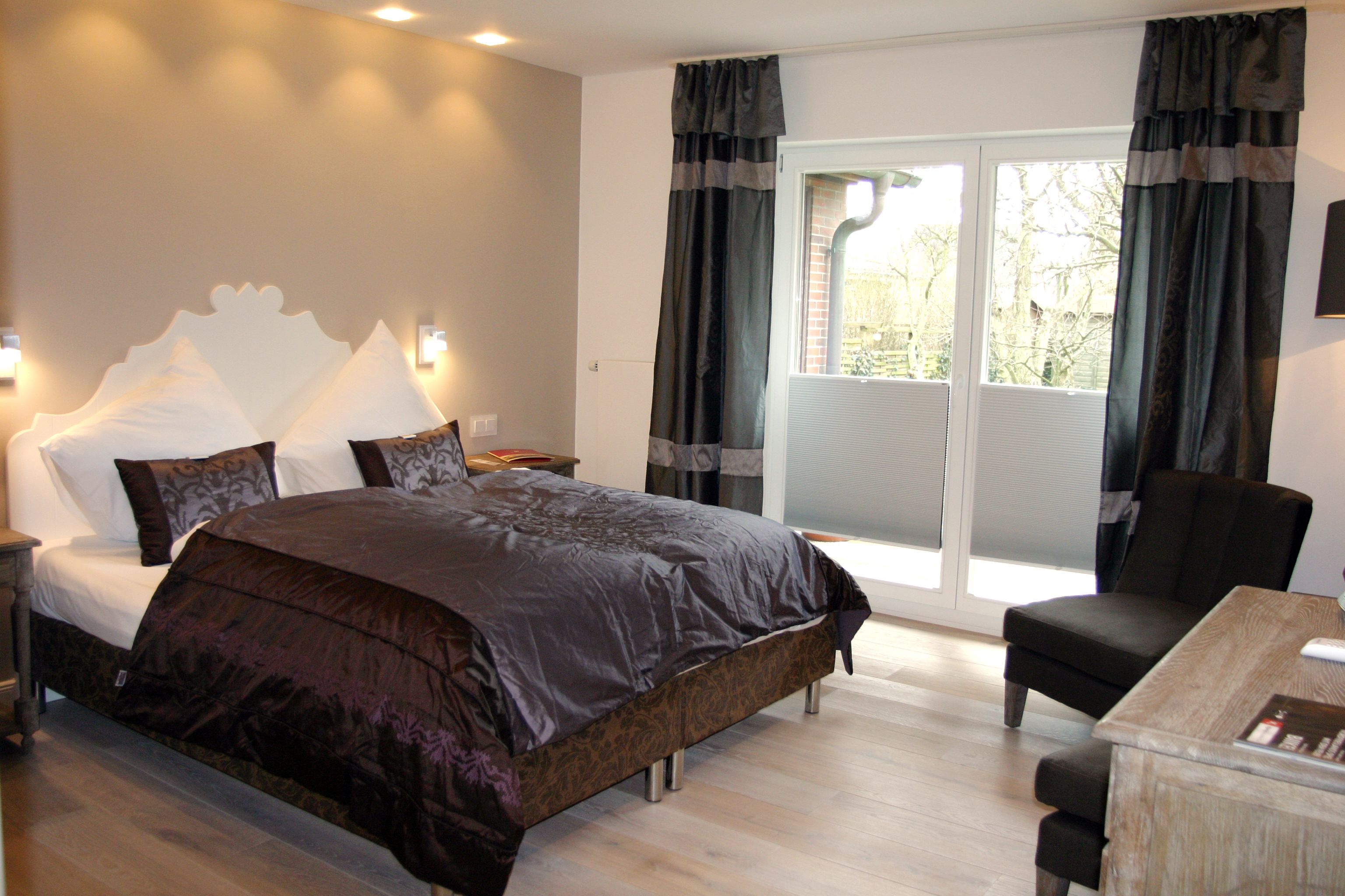 Ein Schlafzimmer mit einem großen Bett, einem Schreibtisch und einem Sessel. Ein Fenster mit Vorhängen bietet Blick auf Bäume.