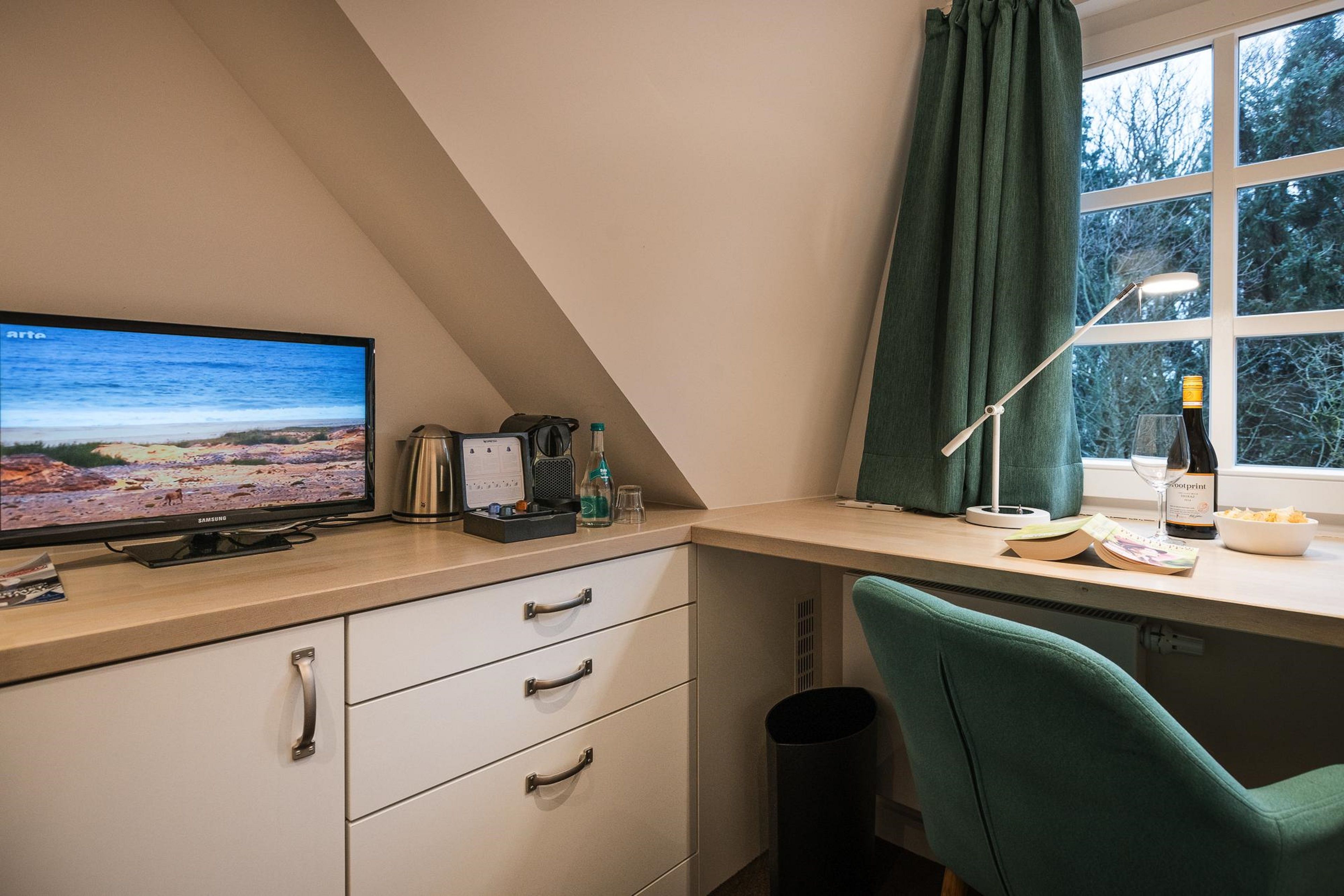 A room with a desk, a chair, a television, a window with curtains, and a trash bin.