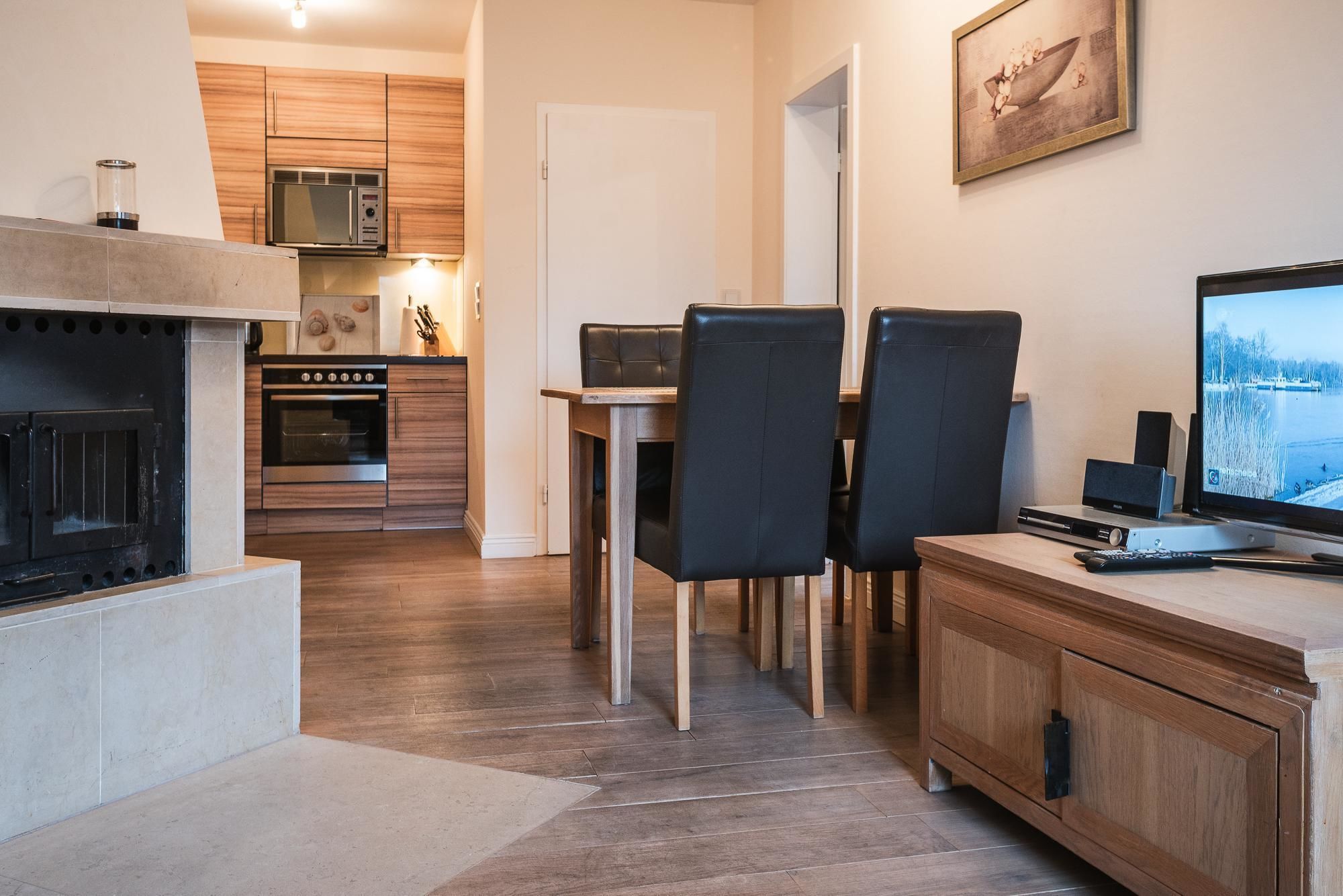 A modern kitchen with wooden cabinets, a built-in oven, and a microwave. There is a dining area with a table and chairs, and a fireplace. A television is mounted on the wall.