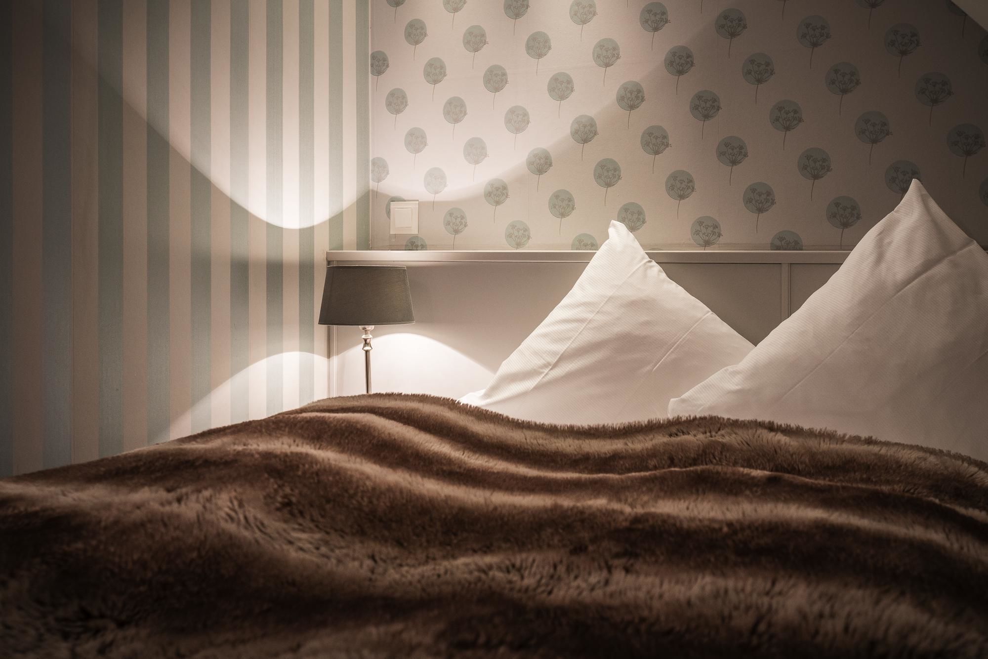 A bedroom with a striped wall, a patterned wallpaper, a bed with white pillows, and a lamp on a bedside table.