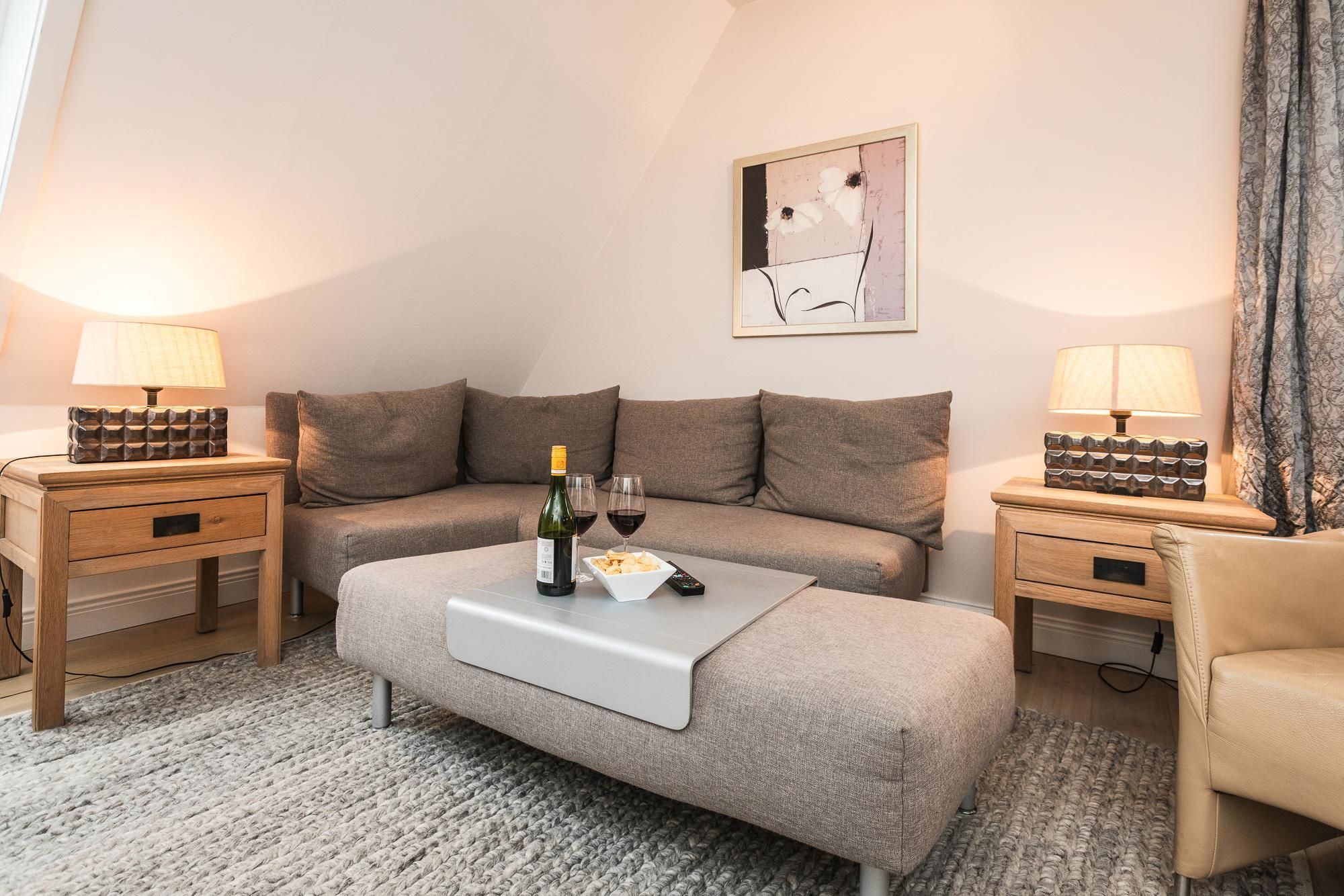 Un salon chaleureux avec un canapé, deux tables de chevet avec des lampes, une table basse sur laquelle se trouve une bouteille de vin, et un tableau encadré au mur.