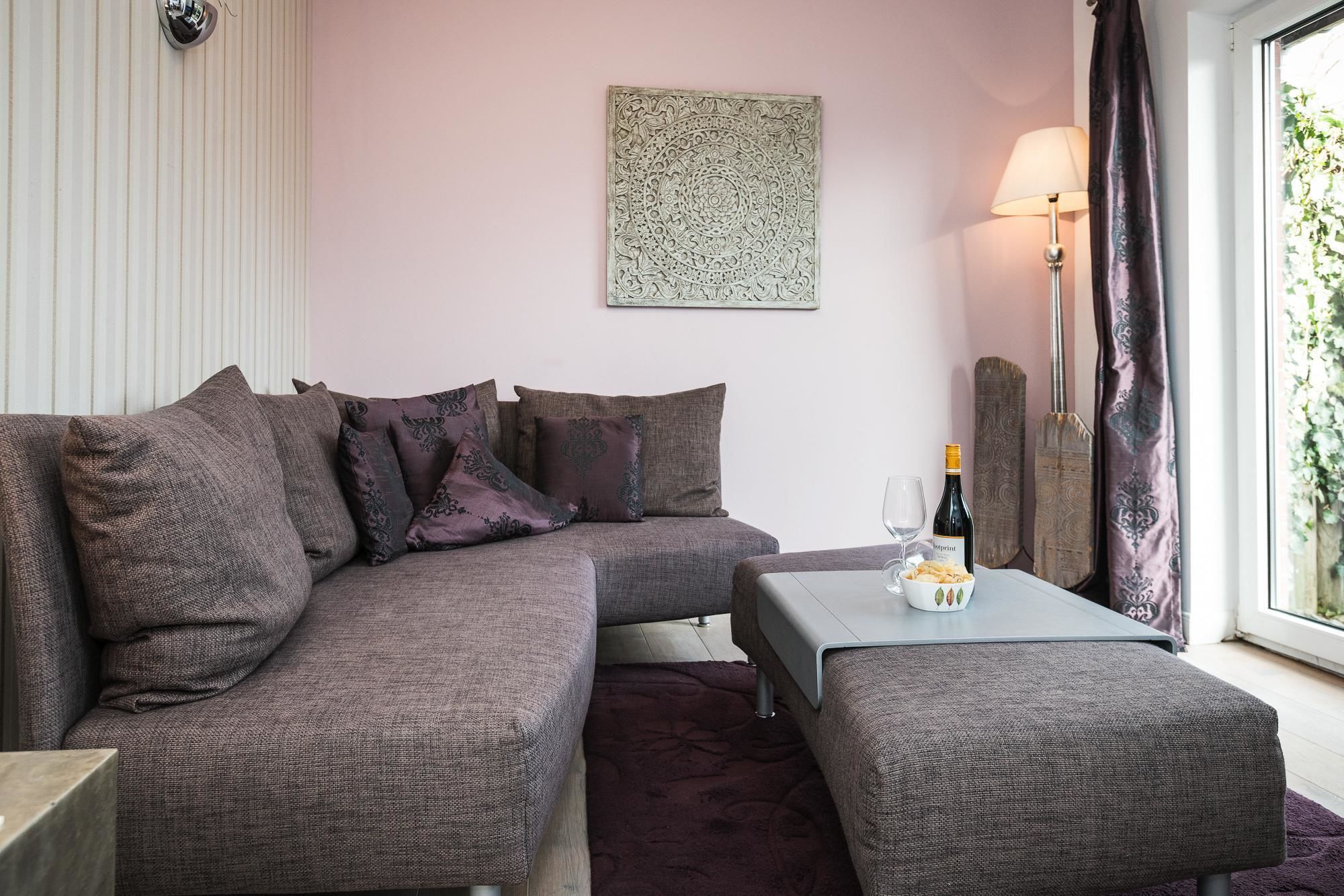Ein modernes Wohnzimmer mit einem Ecksofa, einem kleinen Tisch mit Flasche und Gläsern sowie einem dekorativen Wandbild.