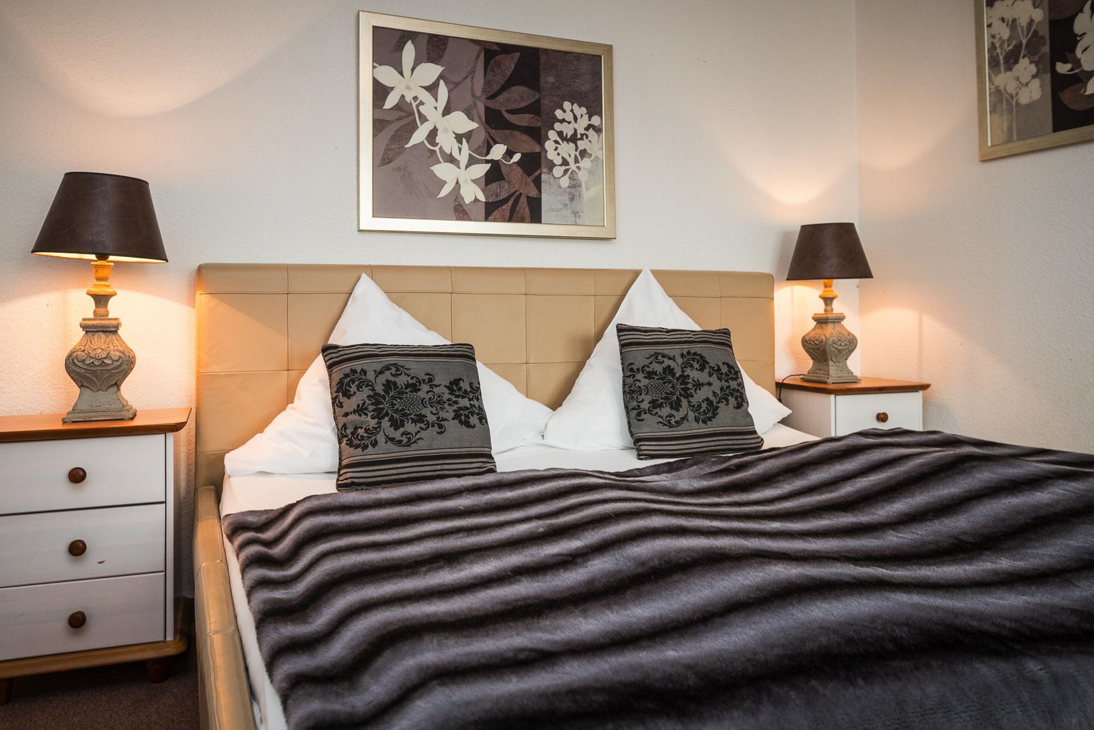 Ein ordentlich bezogenes Bett mit einer gemusterten Decke, zwei Nachttische mit Lampen und ein Blumenbild an der Wand.