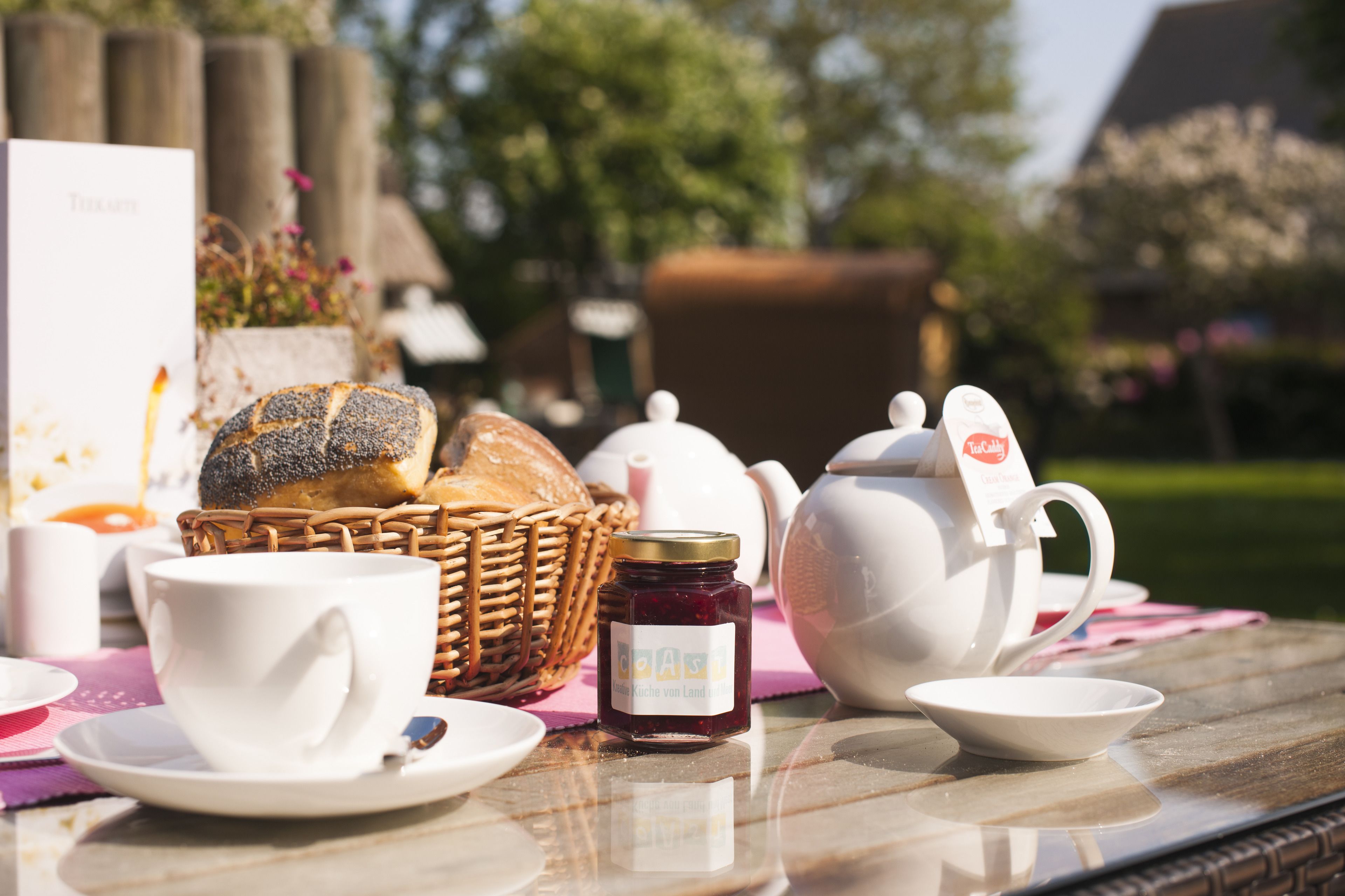 A table set with a teapot, cups, a jar of jam, a loaf of bread, and a basket.