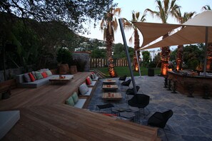 Un patio al aire libre con mobiliario de madera, suelo de piedra y una zona de estar con cojines.