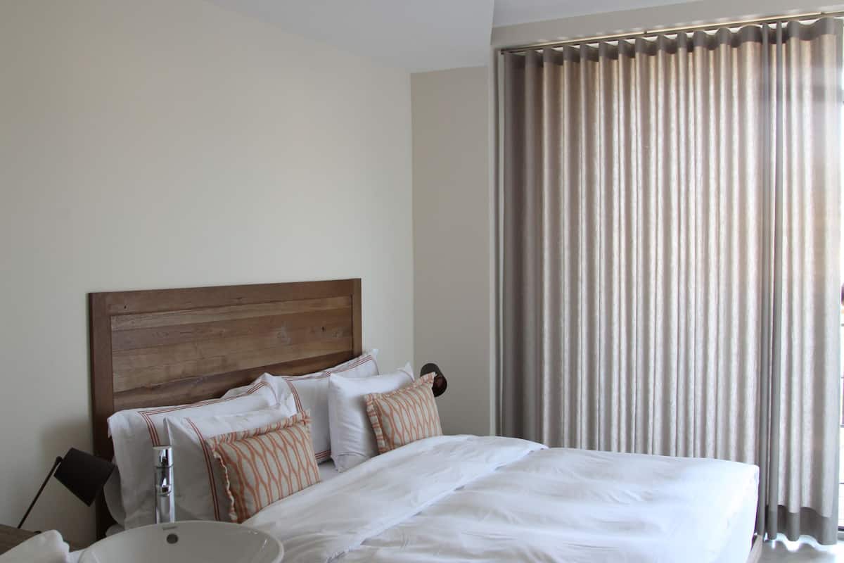 Habitación de hotel con una cama grande, cabecera de madera, mesita de noche con artículos de aseo y vistas al exterior a través de cortinas.