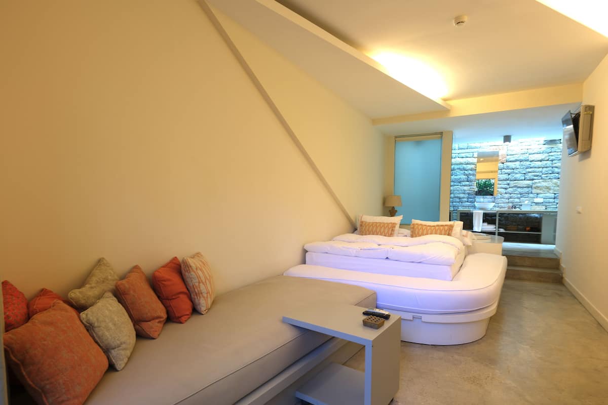 Un dormitorio moderno con cama, sofá y una pequeña mesa. Se observa una pared de piedra y una ventana al fondo.