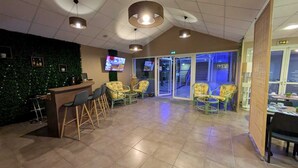 A hotel lobby with a reception desk, bar area, and seating. There are ceiling lights, a green wall with lights, and a TV screen. - Hotel L'Océane Angers Parc Expo (Rives-du-Loir-en-Anjou)