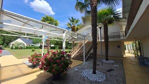 A covered outdoor area with a glass structure, palm trees, and a staircase leading to an upper level. - Hotel L'Océane Angers Parc Expo (Rives-du-Loir-en-Anjou)