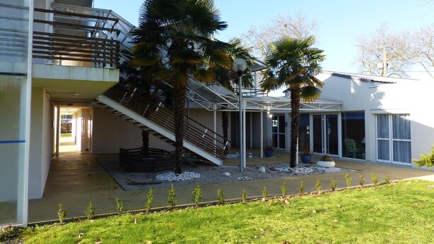 A modern building with a covered outdoor area, palm trees, and a staircase leading to an upper level. - Hotel L'Océane Angers Parc Expo (Rives-du-Loir-en-Anjou)
