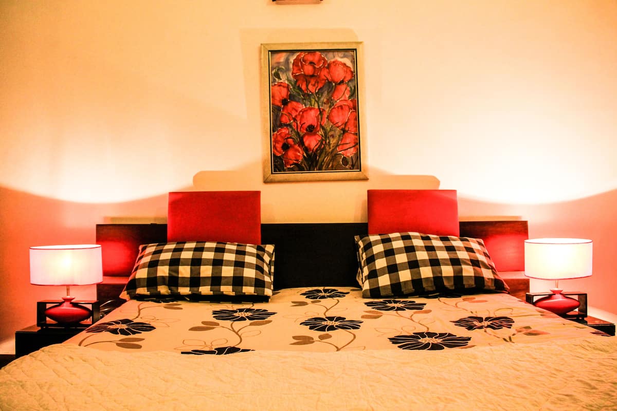 Una cama con cubrecama floral, dos almohadas a cuadros y un cabecero rojo, con un cuadro enmarcado encima.