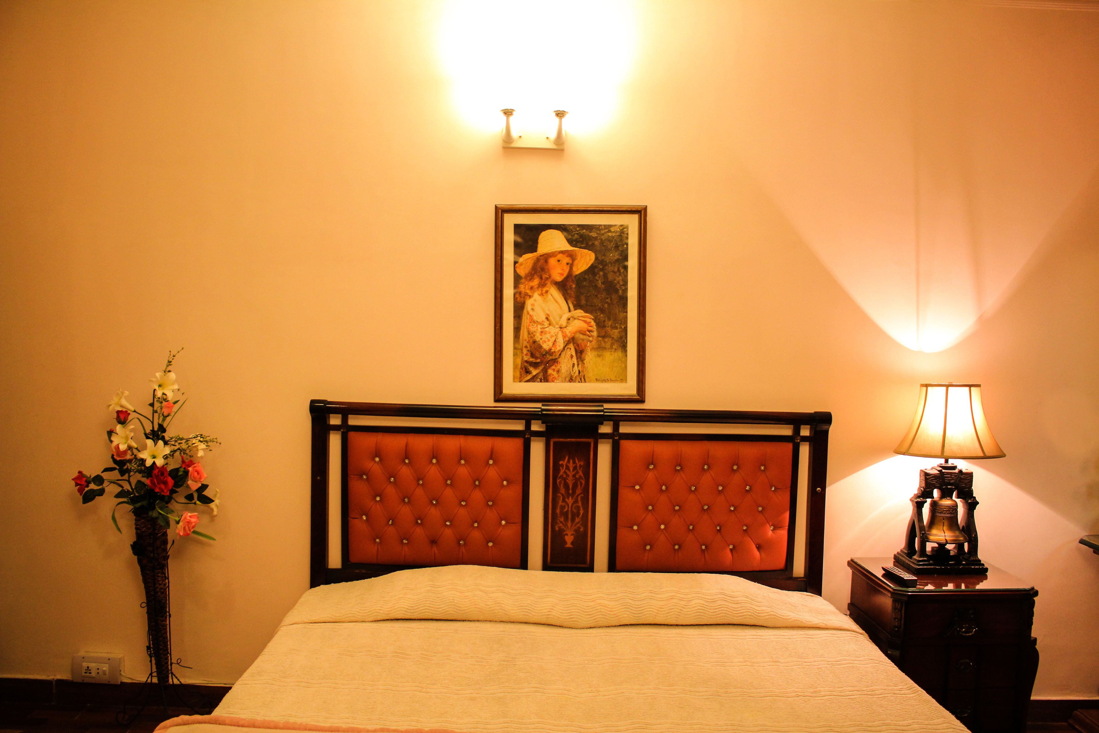 Een hotelkamer met een gestapeld hoofdeinde, een lijst met een portret boven het bed, een bedlamp en een bloemenvaas.