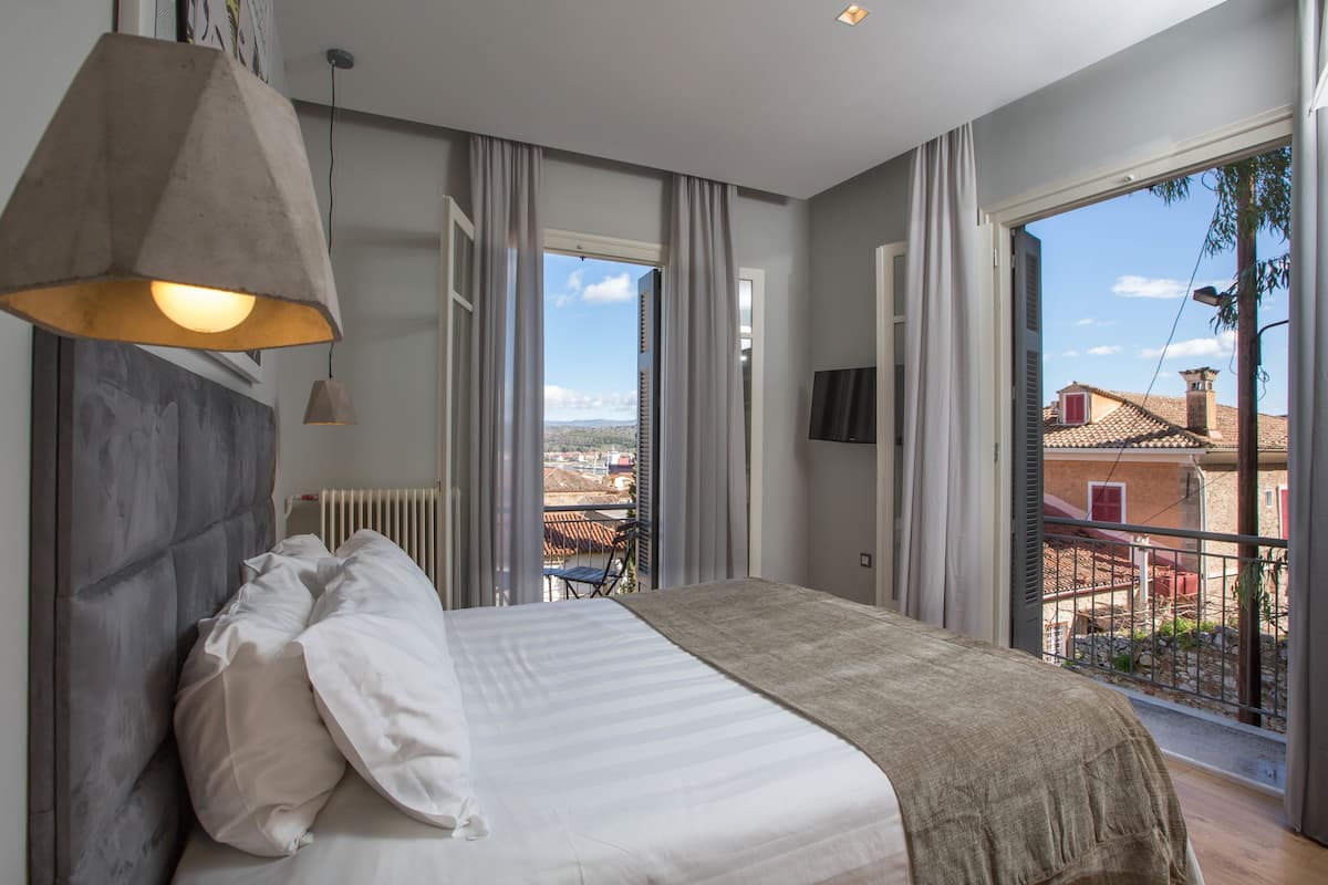 A bedroom with a bed, a hanging light fixture, a radiator, and a view of the city through large windows.