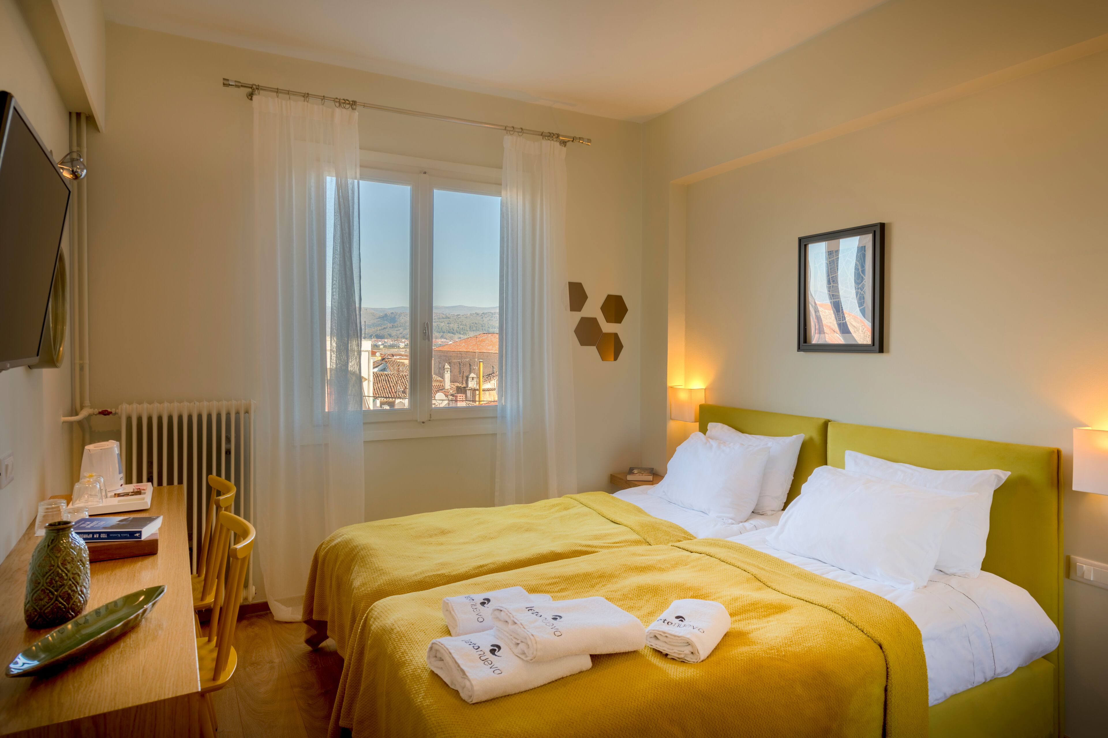 Une chambre d’hôtel avec un lit, une fenêtre avec des rideaux, un radiateur, un bureau avec un vase et des livres, et un mur orné d’un tableau encadré.