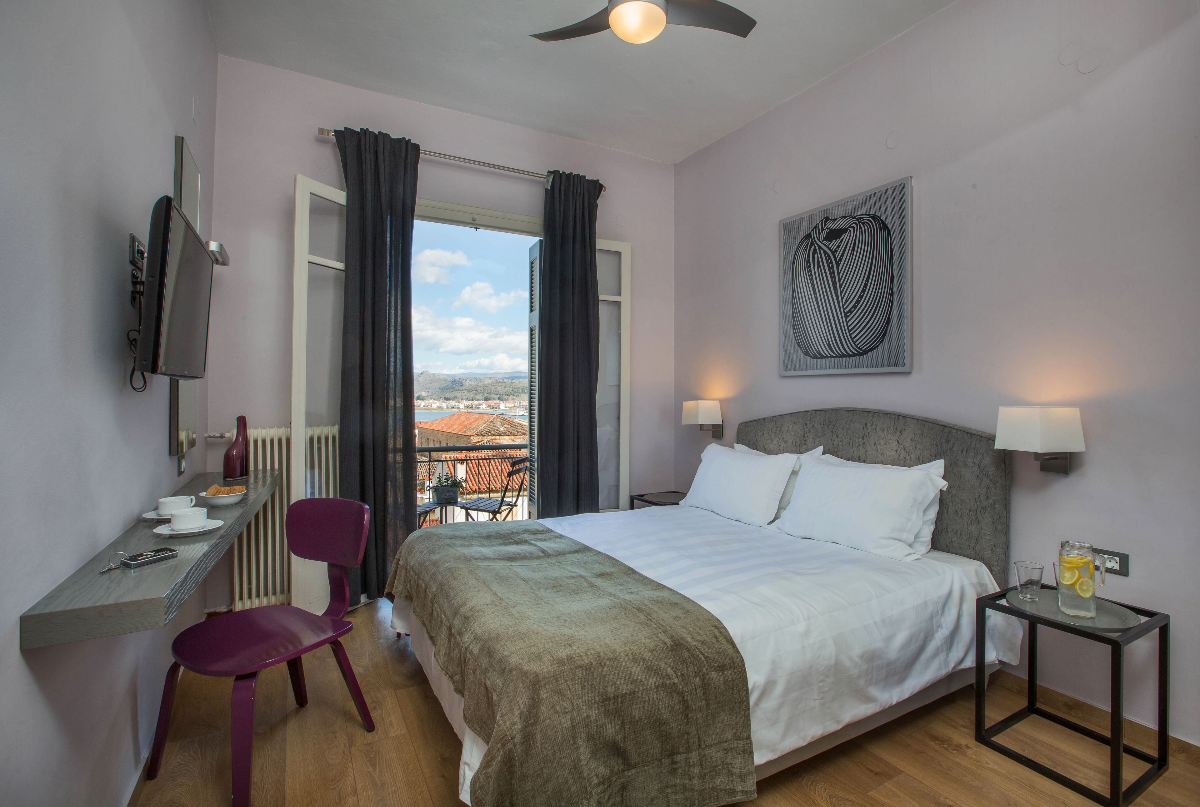 A hotel room with a bed, a desk with a chair, a window with curtains, a wall-mounted TV, a lamp, and a painting.