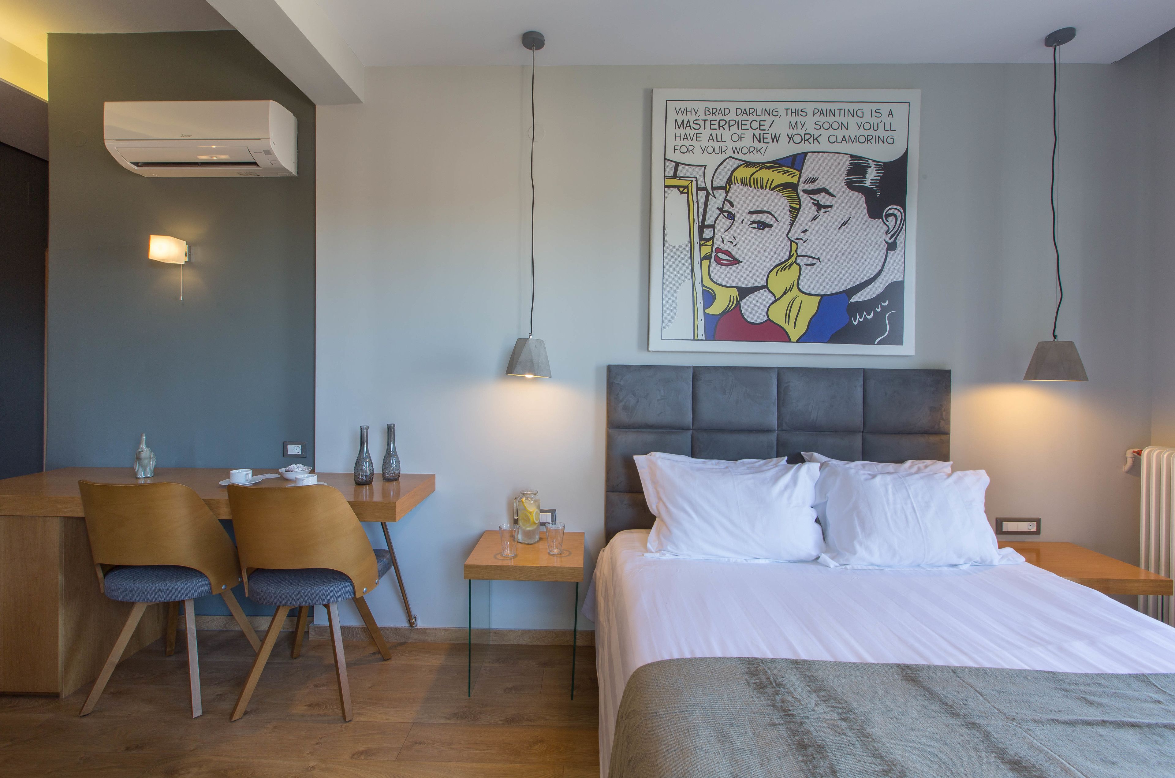 Een hotelkamer met een bed, een bureau met stoelen, een kleine tafel met een lamp en een poster aan de muur.
