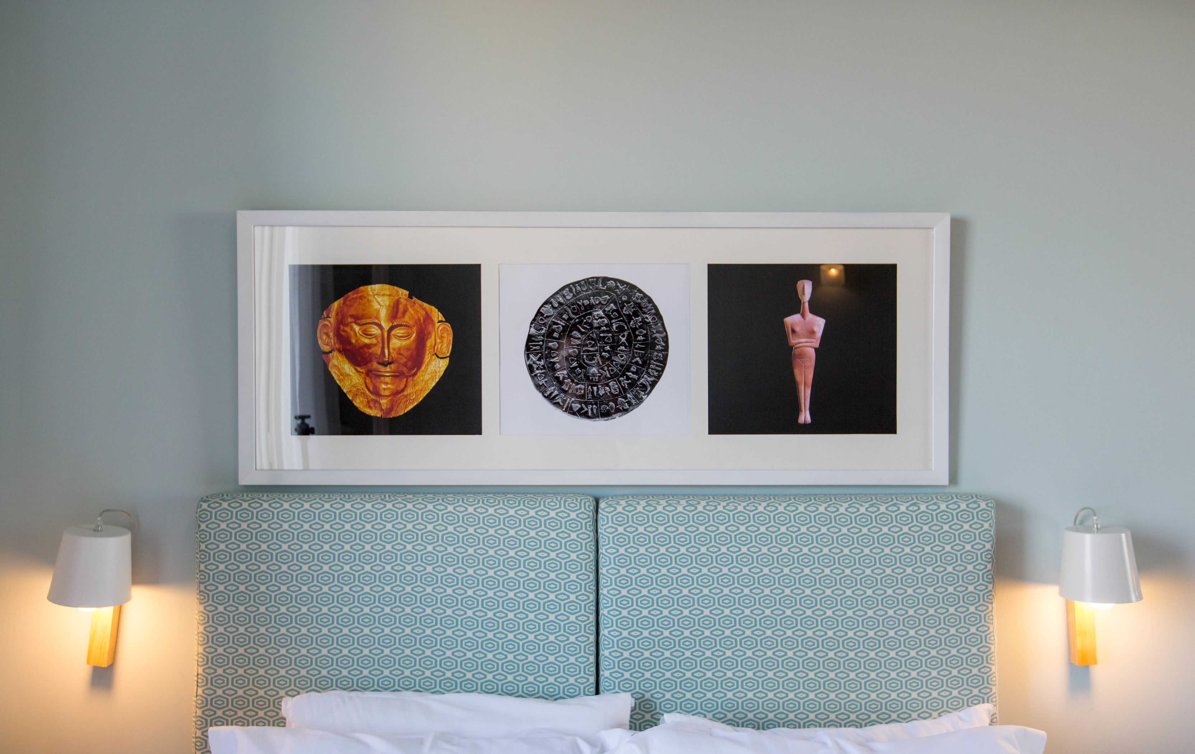 Una obra de arte enmarco sobre una cama con cabecera estampada, dos lámparas fijadas a la pared y un pomo de puerta visible.