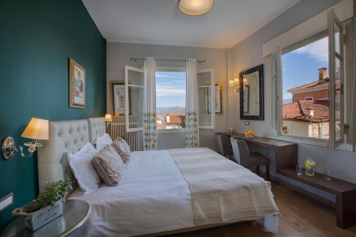 A bedroom with a bed, bedside table, lamp, and a view of the city through open curtains.