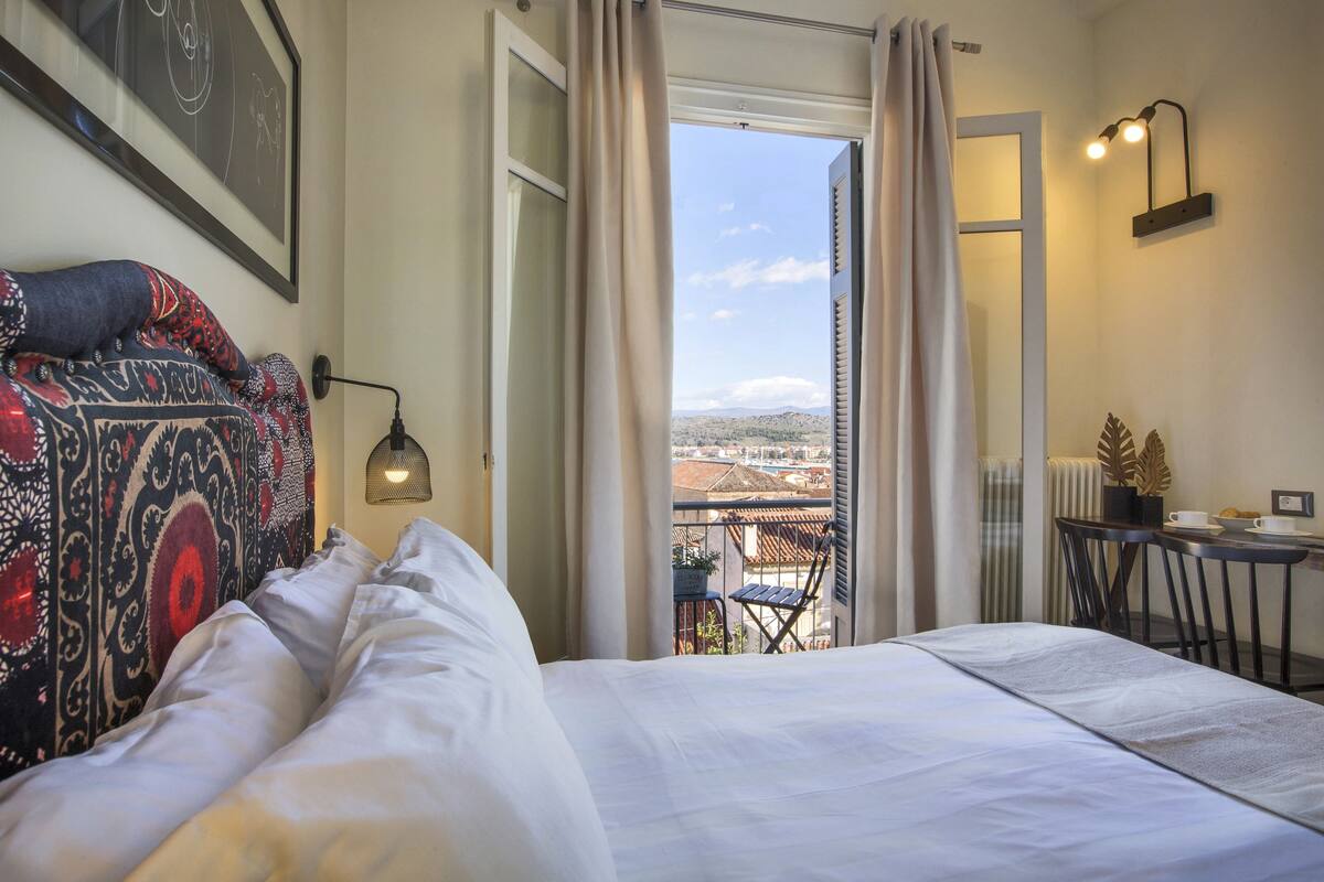 A hotel room with a bed, a view of the city, a small balcony, and a table with a chair.
