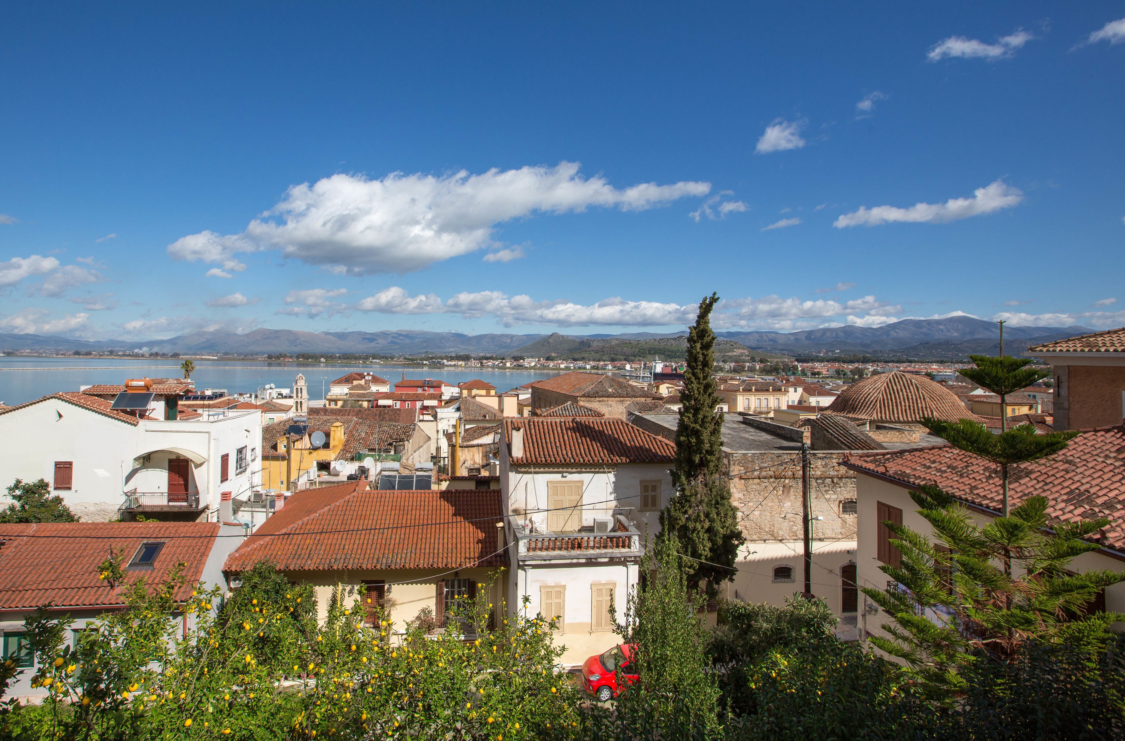Un pueblo costero con edificios de techos de teja rojiza, cielo azul despejado y vistas al mar y montañas a lo lejos.
