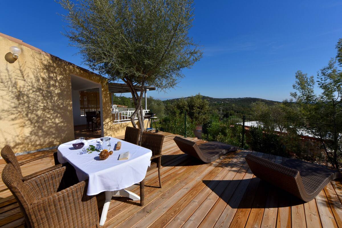 An outdoor dining area with a table set for two, surrounded by wicker furniture, under a pergola with a view of the countryside.