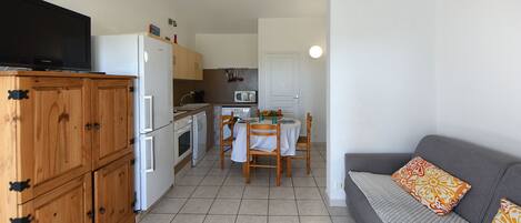 Et lille køkken med køleskab, mikrobølgeovn og komfur. Spisebord med stole og sofa med pyntepuder.