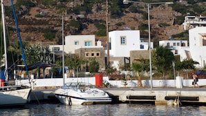 A coastal marina with boats docked, white buildings with blue accents, and a hill with a tower in the background. - Villa Patmos Netia (Patmos)