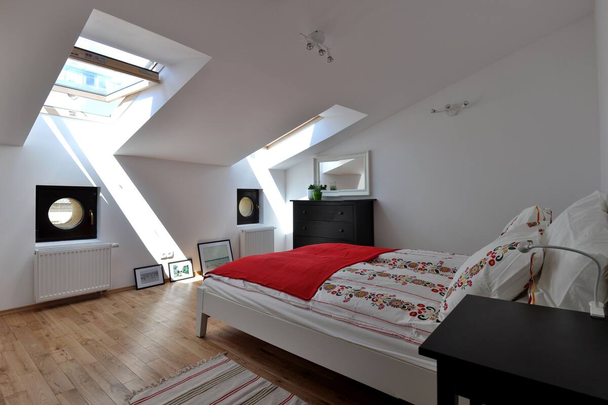Un dormitorio con tragaluces, una cama con una colcha roja, un aparador y un espejo en la pared.