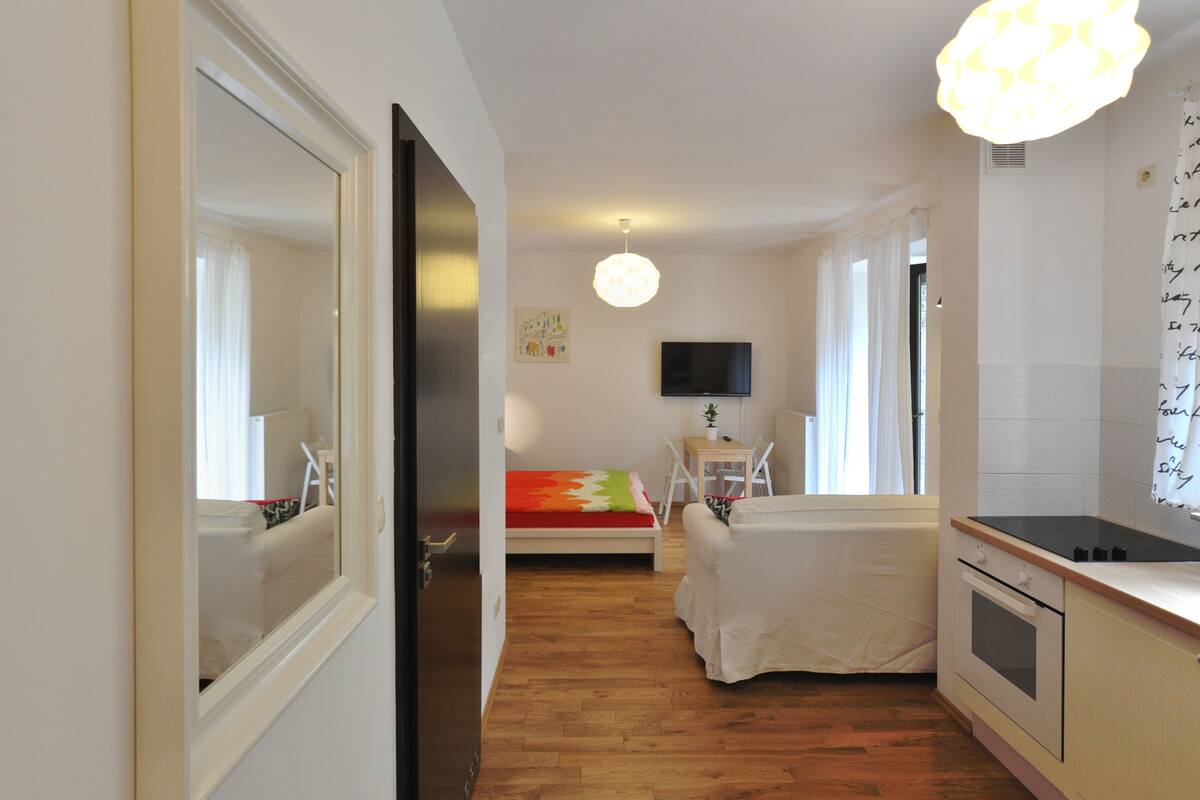 Una habitación de hotel con piso de madera, una cama con una colcha colorida, una pequeña mesa con una silla, un televisor montado en la pared y una zona de cocina con estufa y horno.