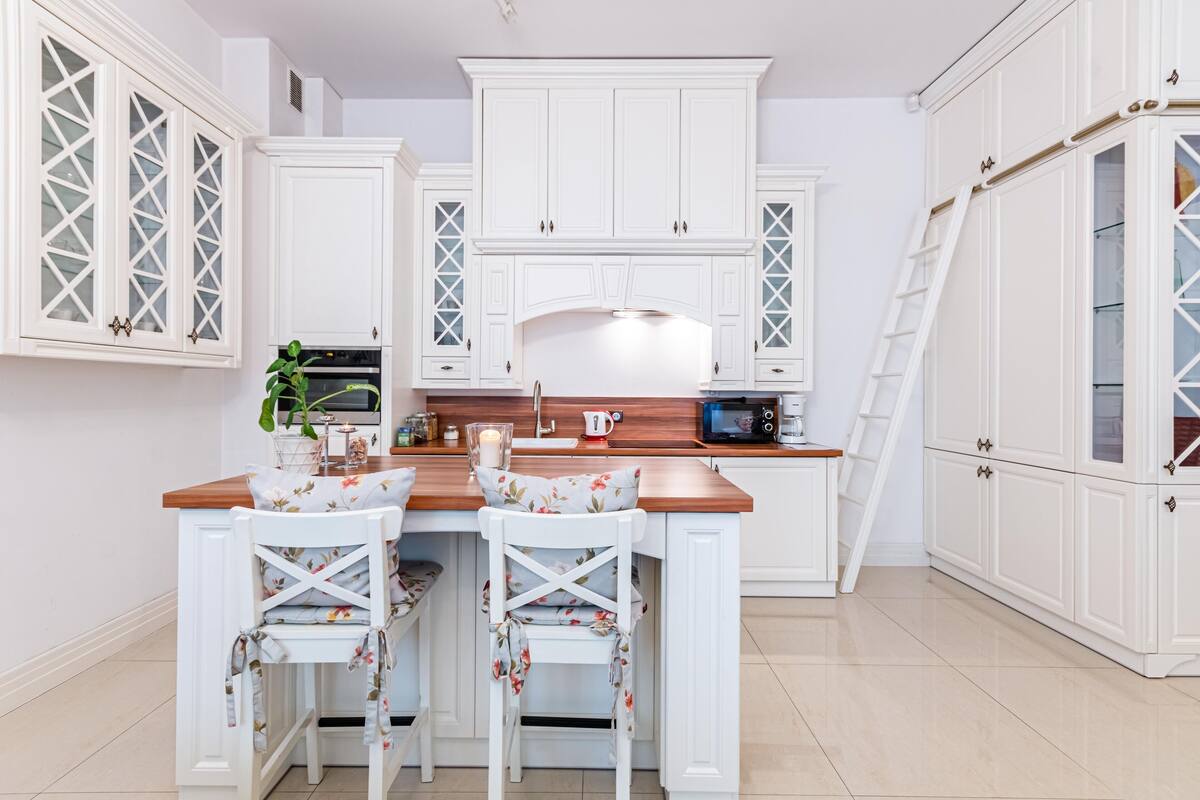 Una cocina con armarios blancos, encimera de madera y dos sillas blancas con tapicería floral.