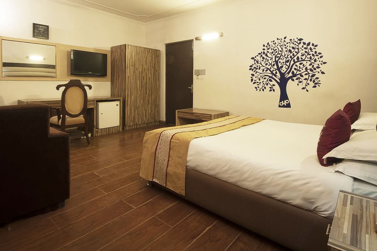 Habitación de hotel con cama, escritorio, silla, televisor y un cuadro de un árbol.