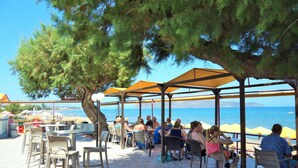 An outdoor seating area with tables and chairs under a shaded structure, overlooking a body of water. - Sea Side Apartments (Chania)