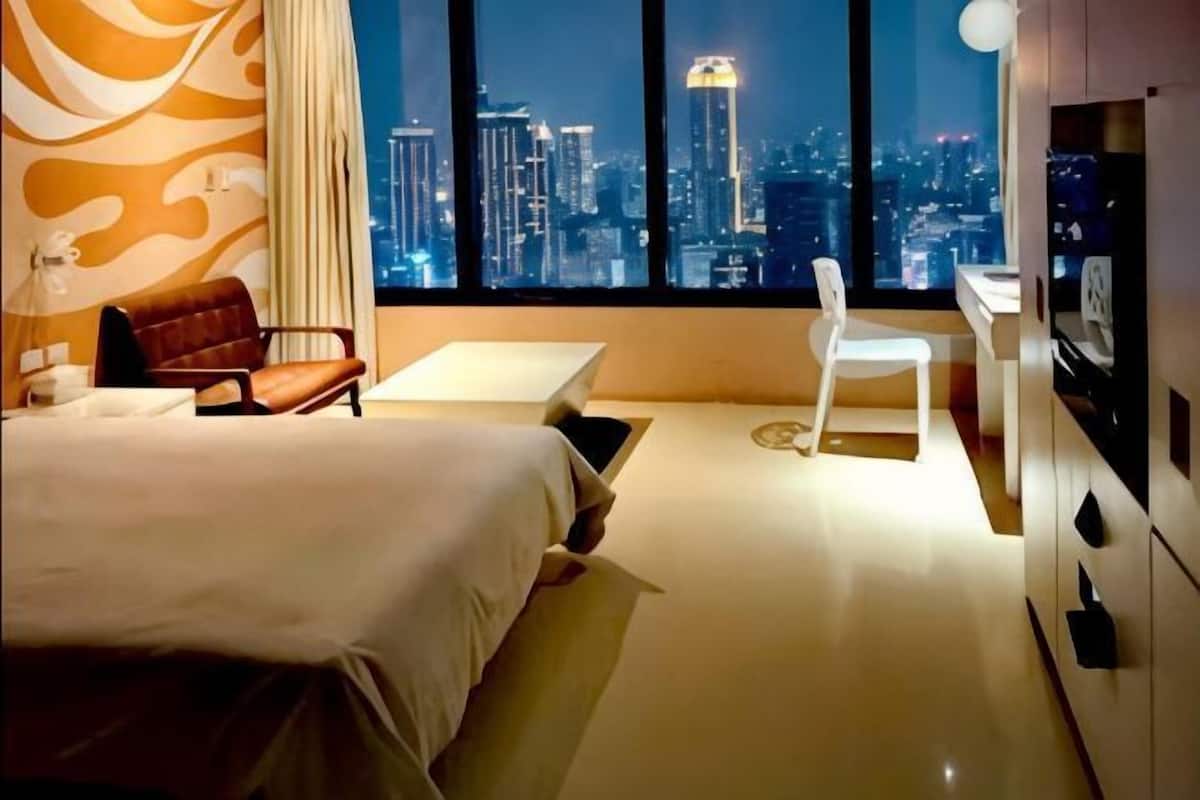 A hotel room with a bed, a desk, a chair, and a view of a cityscape at night.