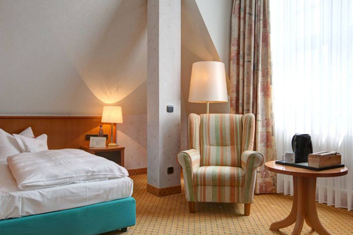 A hotel room with a bed, a striped armchair, a nightstand with a lamp, a small table with a tray, and a window with curtains.