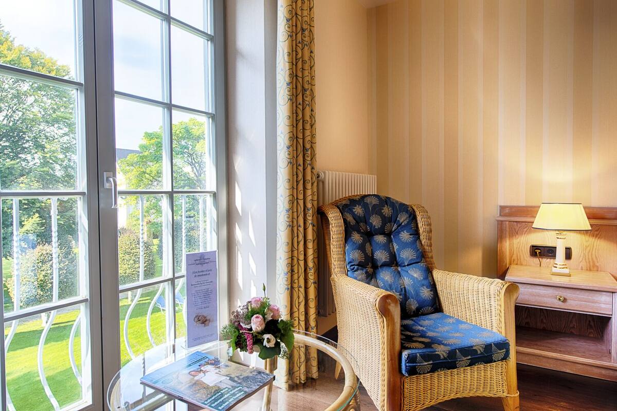 A room with a wicker chair, a table with a vase of flowers, and a nightstand with a lamp.