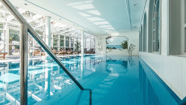 Una piscina interna con acqua cristallina, lettini prendisole e vista sull'oceano attraverso ampie finestre.