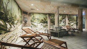 Un'area benessere con lettini di legno, una fontana in pietra e un murale raffigurante un paesaggio tropicale.