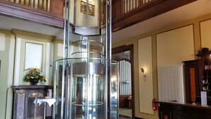 Una hall con un ascensore centrale in vetro, una reception e un vaso decorativo con fiori.