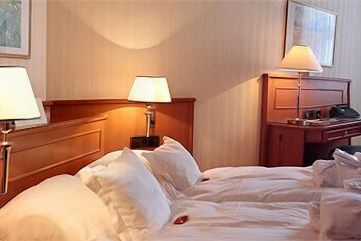 A hotel room with a bed, bedside lamps, a desk, a chair, and a small table with a lamp.