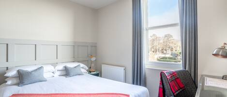 Habitación de hotel con una cama grande, un escritorio con lámpara y vistas a árboles por la ventana.