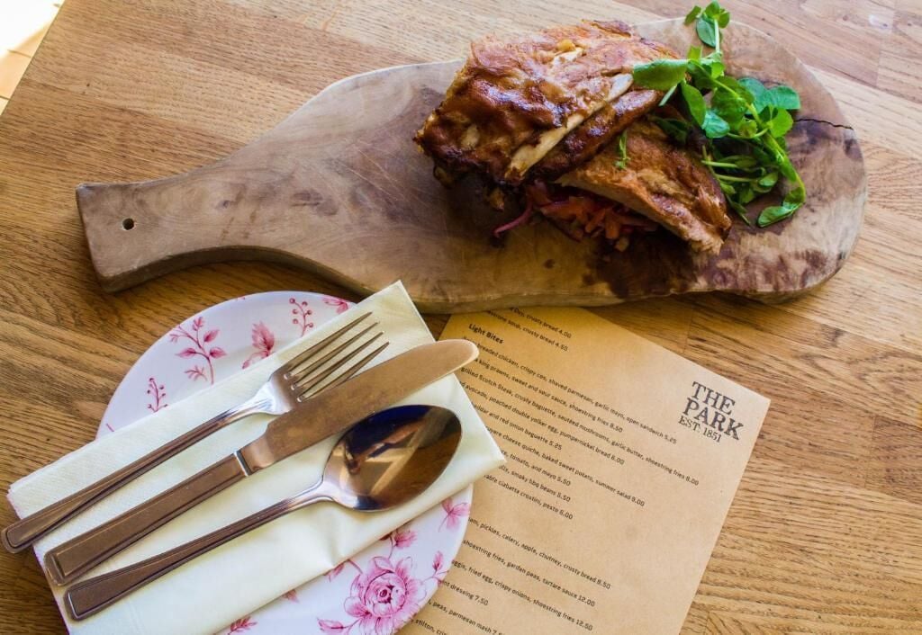 A wooden serving board with a sandwich and greens, a floral plate with cutlery, and a menu.