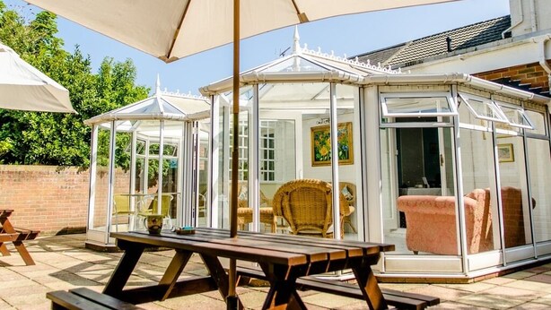 A glass conservatory with a wooden table and chairs, surrounded by a brick wall and trees. - The Avenue (Shanklin)