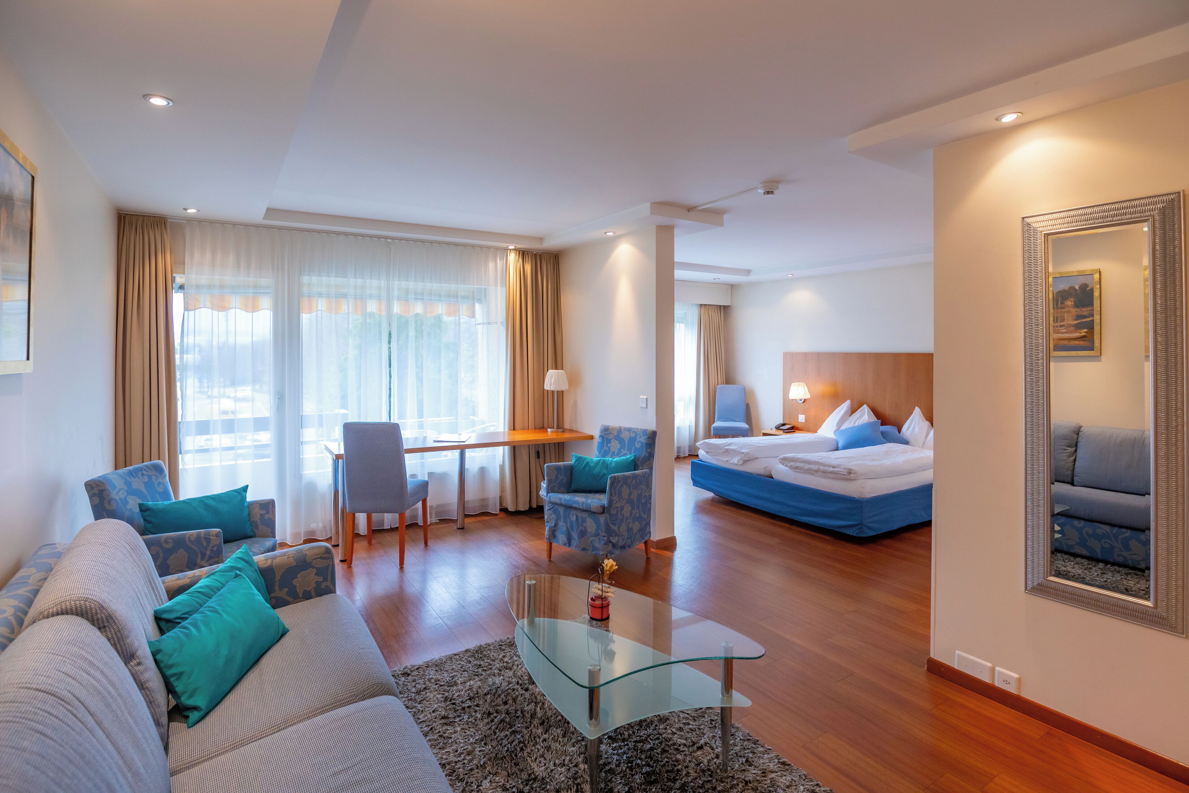 Quarto de hotel moderno com sofá, poltrona, mesa de centro de vidro e uma cama.