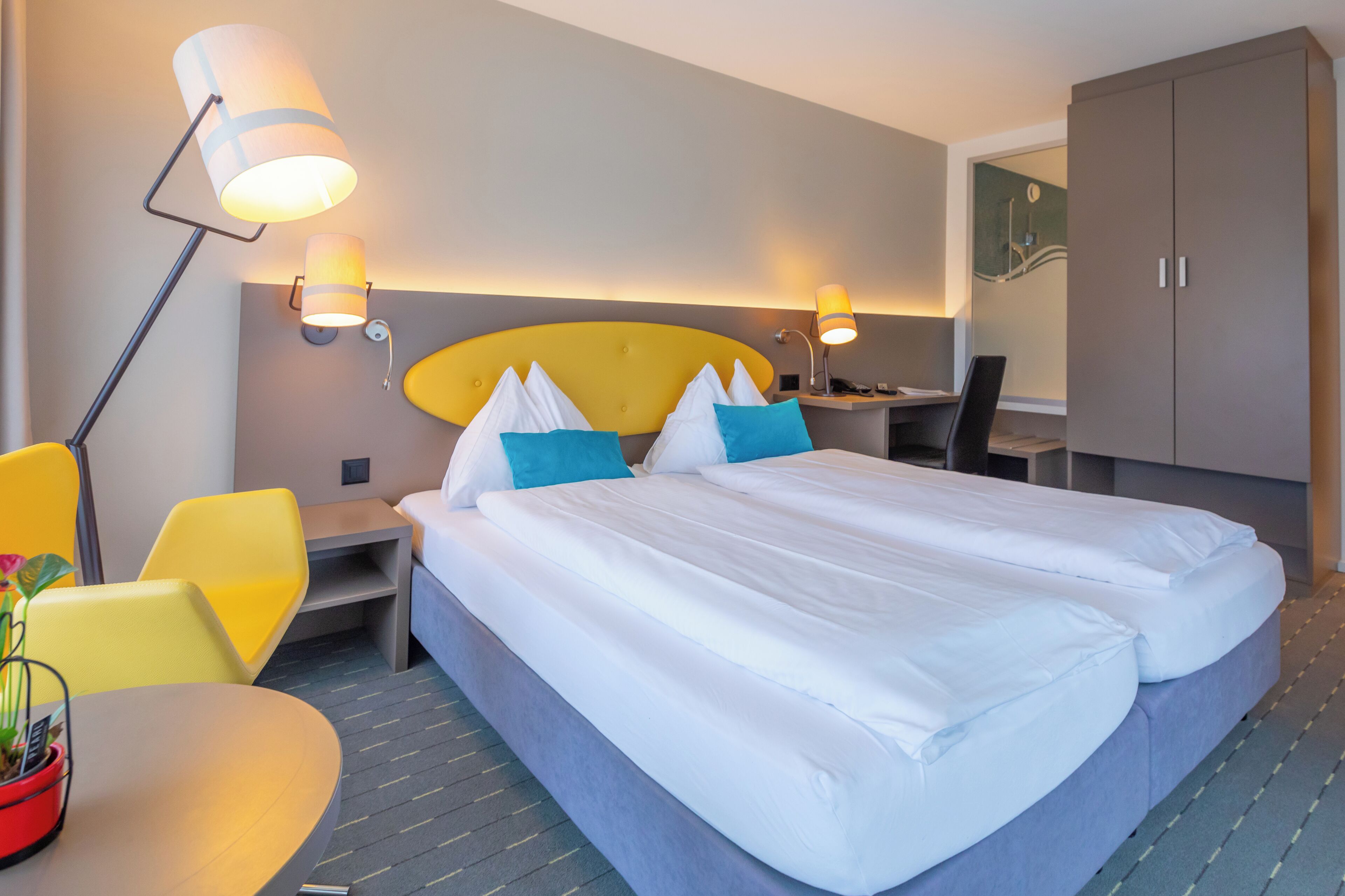 A hotel room with a double bed, two bedside lamps, a desk with a chair, and a wardrobe.