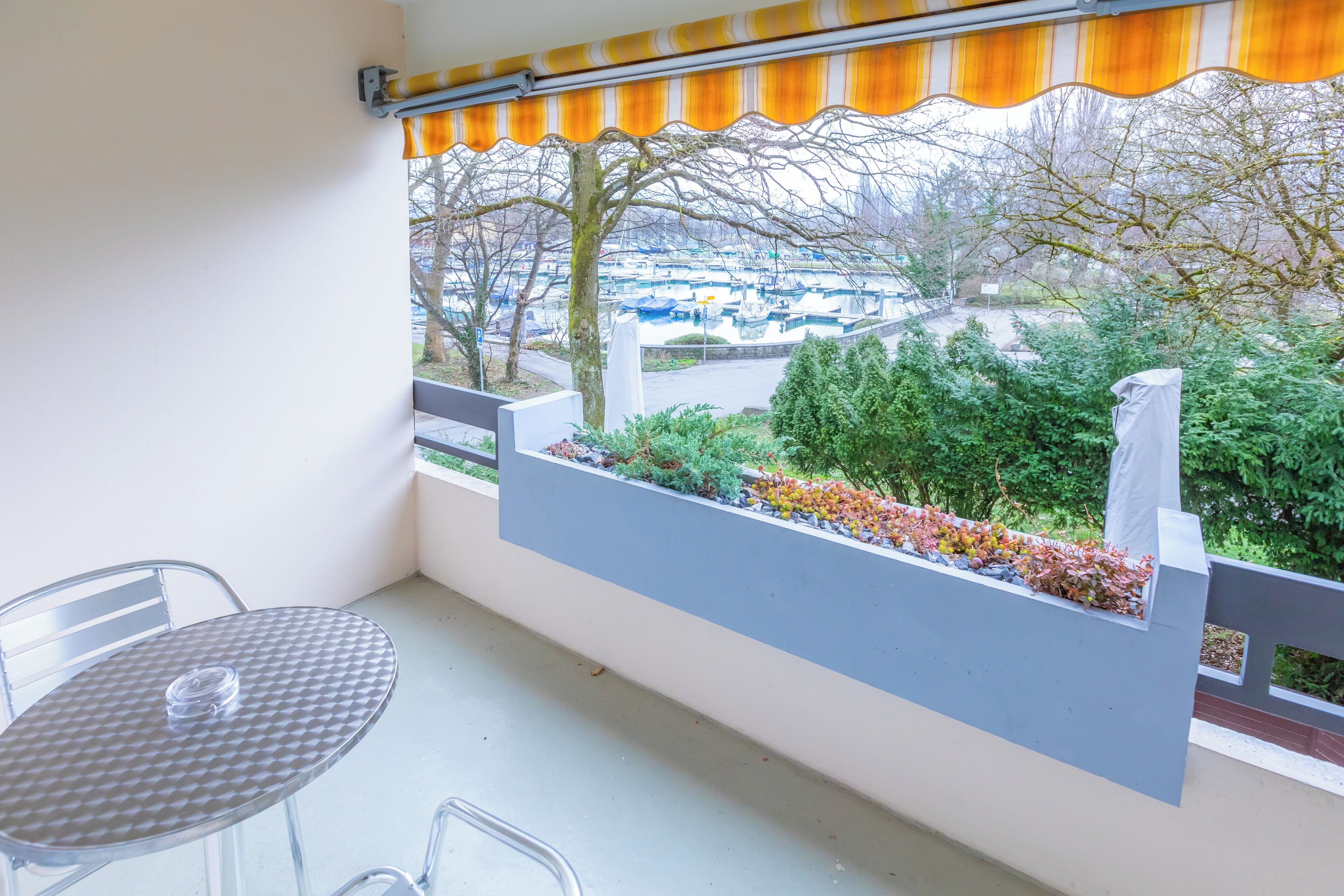 A balcony with a table and chairs, a view of a parking lot, and a row of trees.