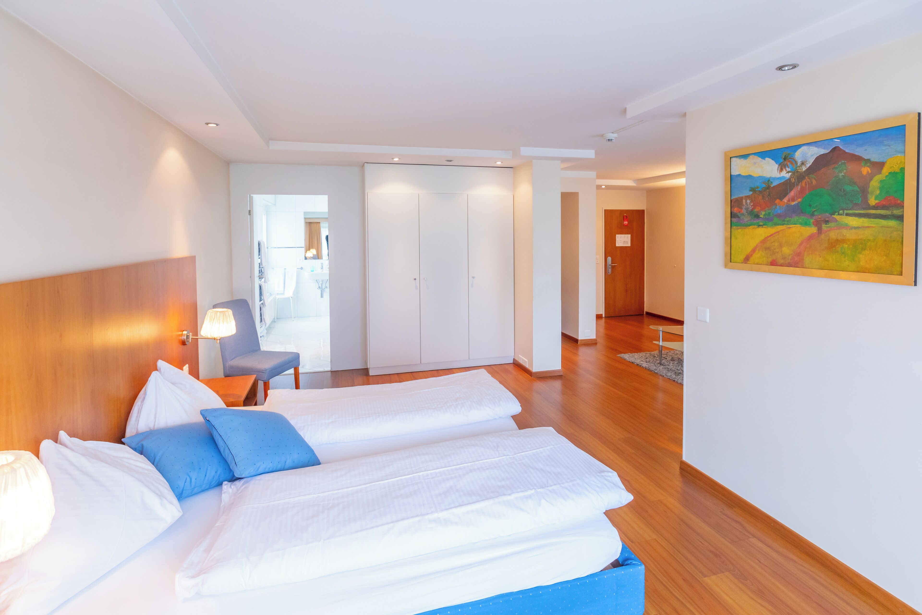 A hotel room with a bed, a chair, a desk, and a painting on the wall.