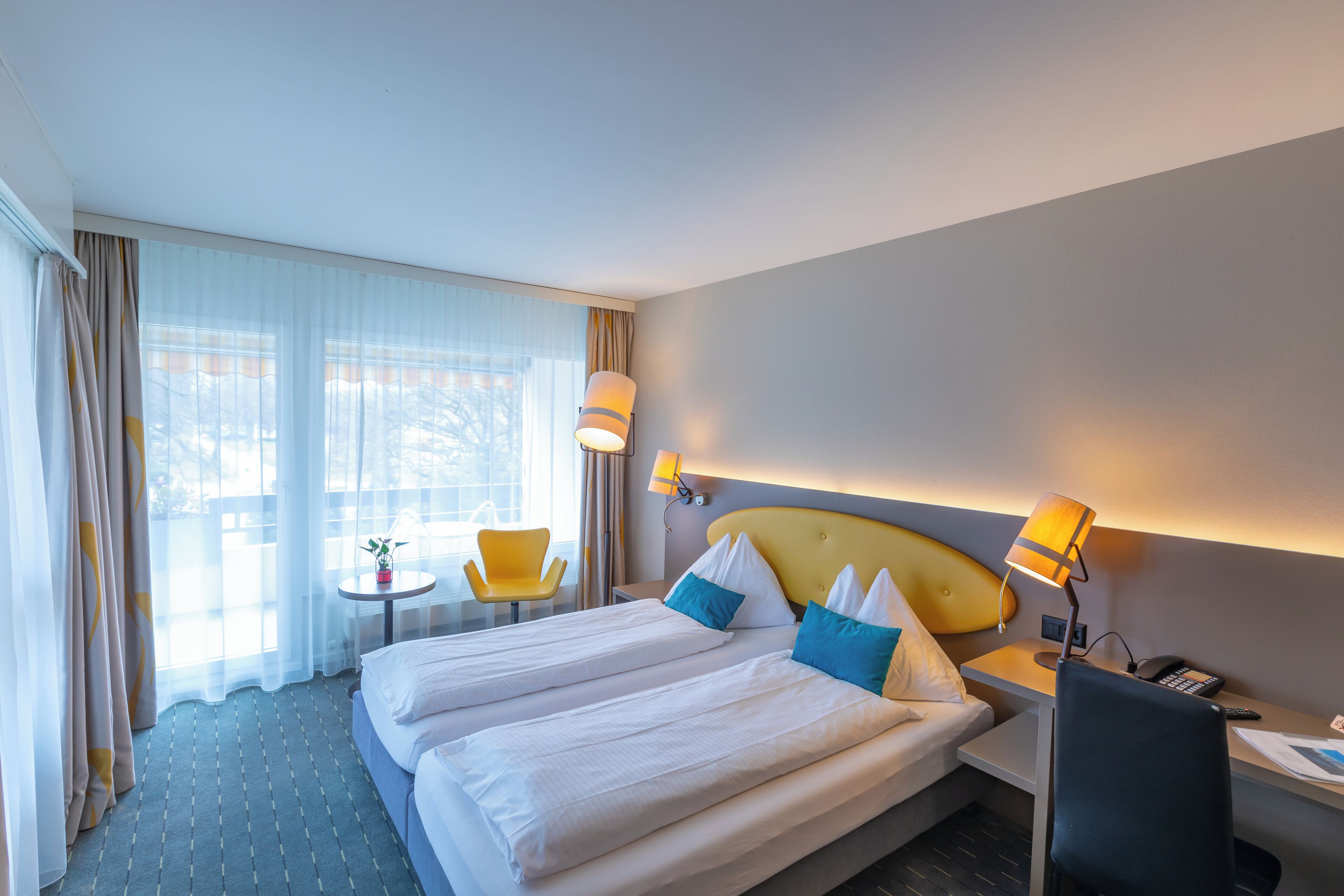 A hotel room with a large bed, two bedside tables, a desk with a chair, a small table with a chair, and a window with curtains.