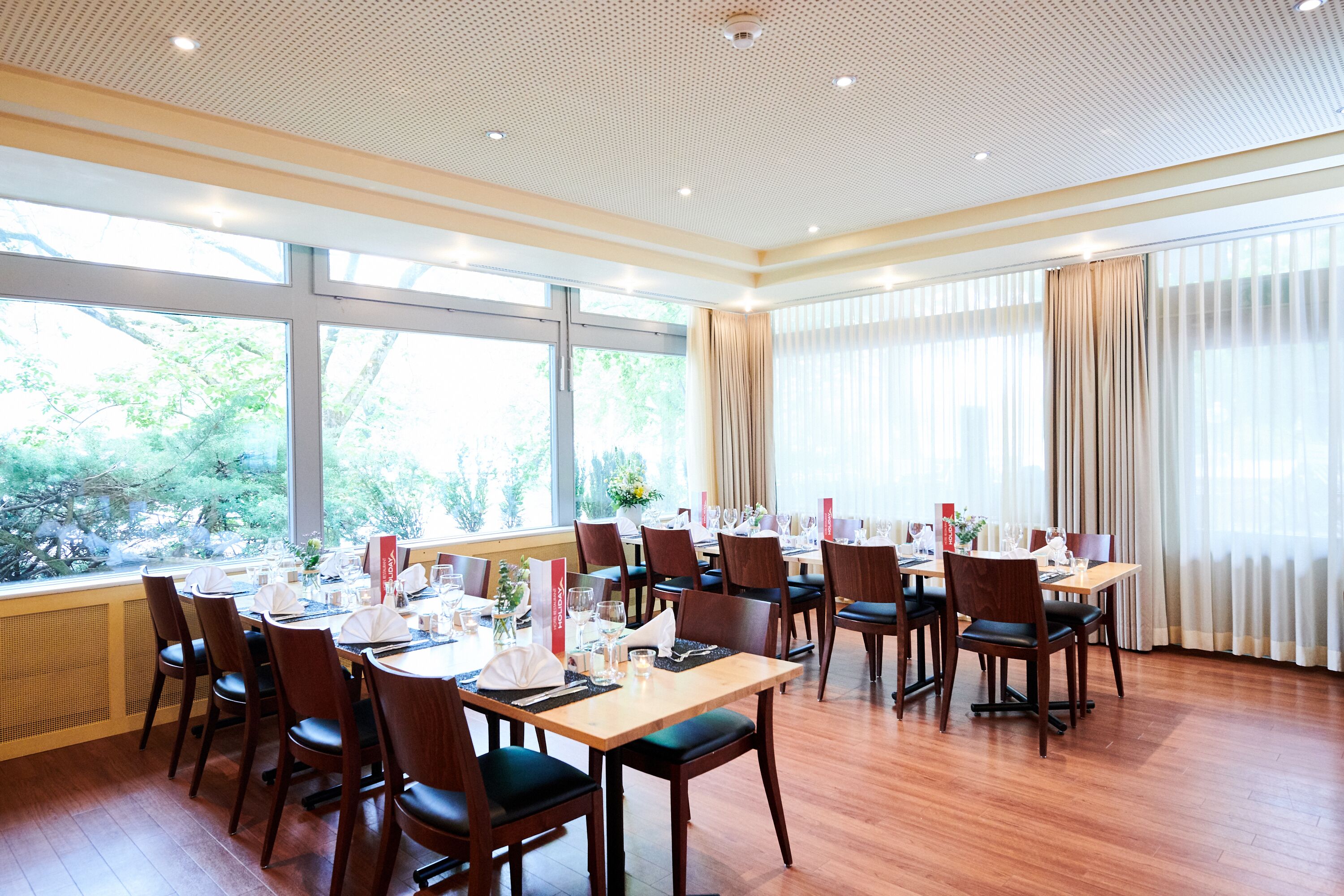 A dining area with multiple tables set for a meal, large windows with curtains, and a ceiling with a grid pattern.