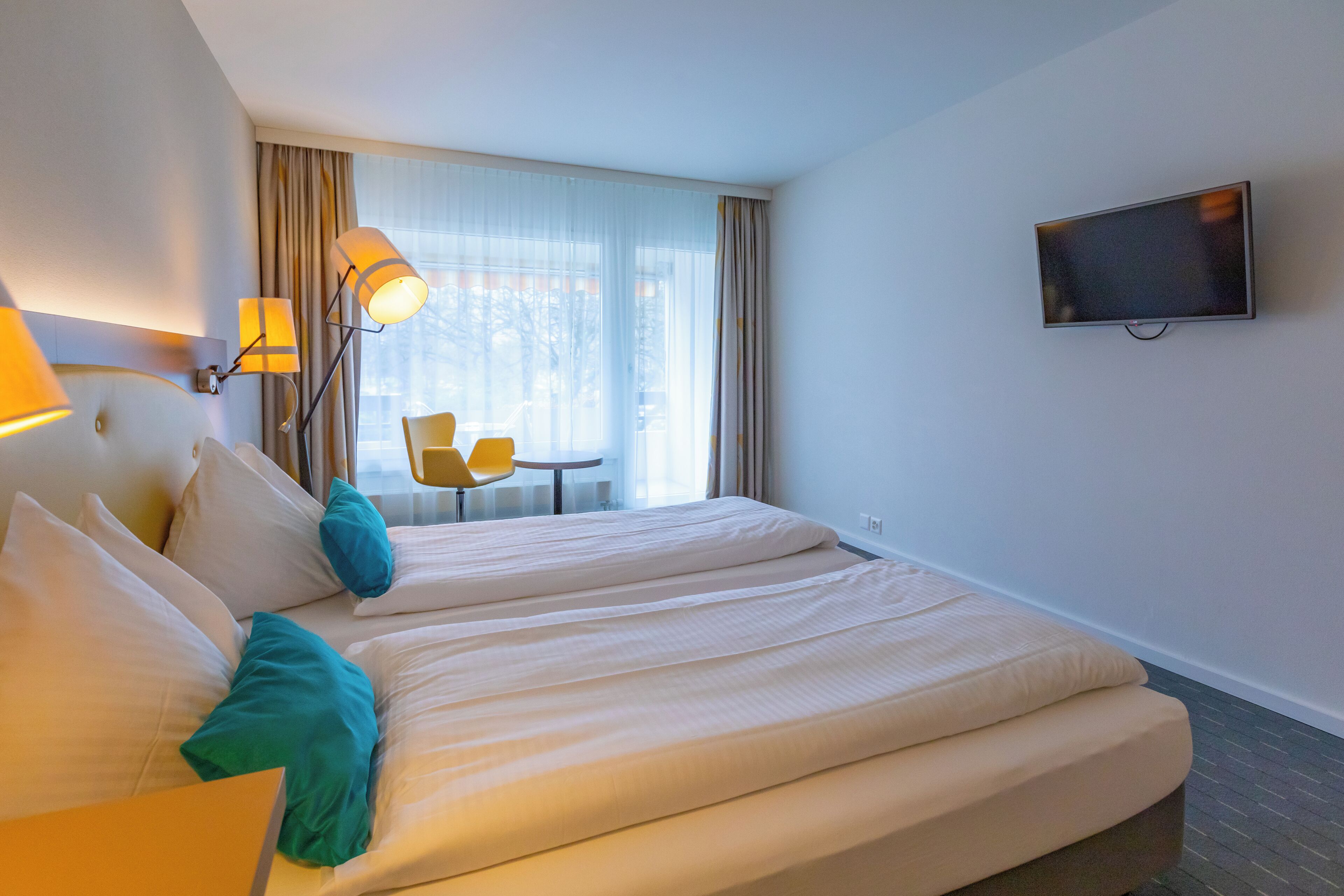 Habitación de hotel con dos camas, televisor en la pared, escritorio con silla y ventana con cortinas.