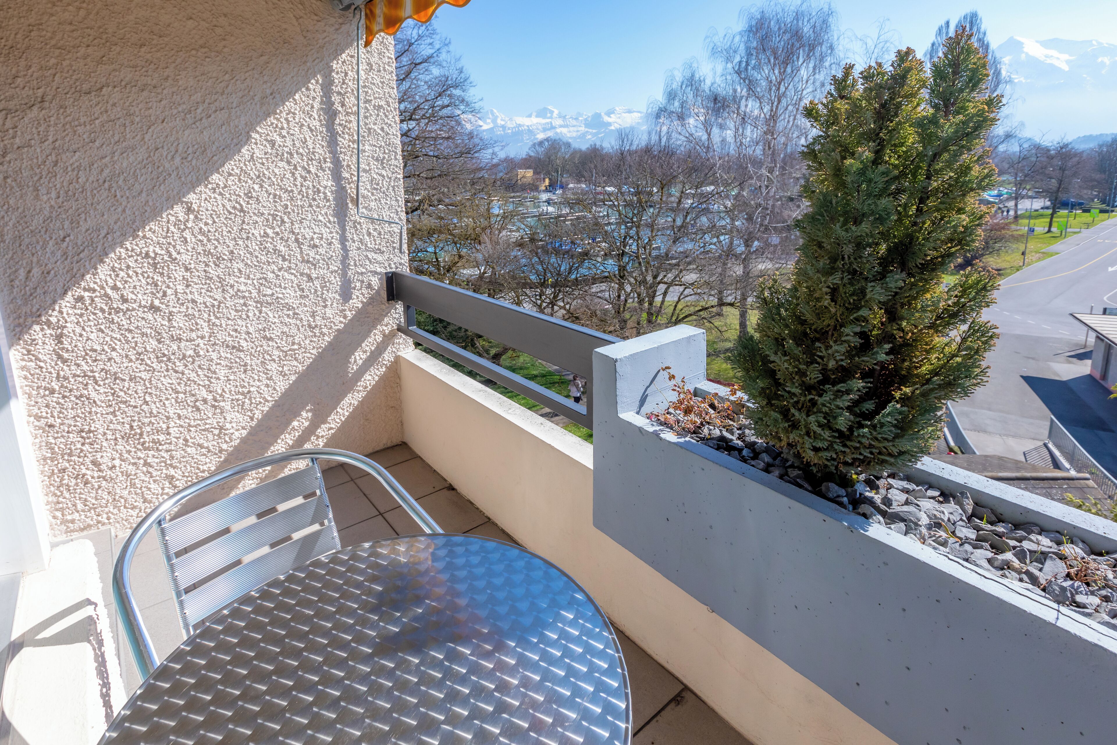 Un balcon avec une table et des chaises en métal, un petit arbre, et une vue sur une route et des montagnes en arrière-plan.